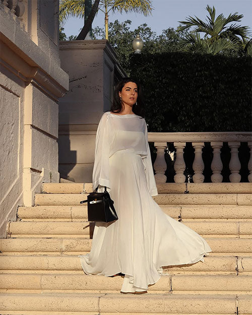 A person is walking down a set of wide stone steps in an outdoor setting, holding a structured black handbag in the left hand. The outfit is a flowing, floor-length white dress with long, loose sleeves and a soft, draped silhouette that moves elegantly with the steps. The background features a classic architectural design with light-colored stone balustrades and walls, and there is greenery visible behind the railing. 
