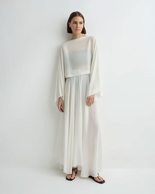 A person is standing against a plain white background wearing a flowing, floor-length white dress. The dress features a sheer, loose-fitting top with wide, draped sleeves and a slightly cropped hem that overlaps a high-waisted, softly gathered skirt. The fabric appears lightweight and semi-transparent, creating an airy and elegant look. The outfit is paired with simple black flat sandals, adding a minimalist touch to the overall style.