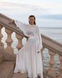 A person is leaning against a stone balustrade overlooking the ocean, wearing a flowing white outfit. The ensemble consists of a sheer, loose-fitting top with wide sleeves and a high-waisted, floor-length skirt made of lightweight fabric. The setting features stone steps and railings in the foreground, with calm water and a cloudy sky in the background.