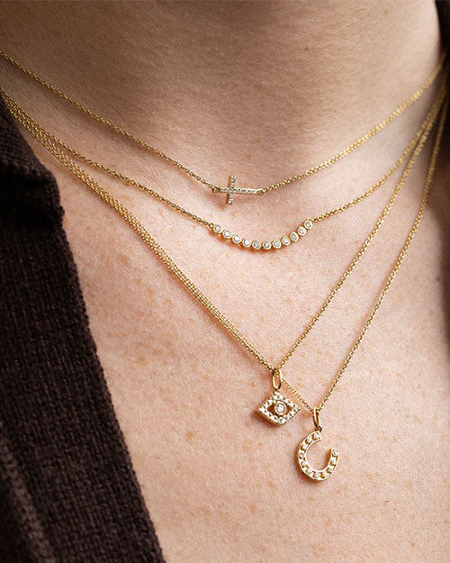 Close-up of layered gold necklaces featuring four different pendants: a horizontal cross with small gemstones, a curved bar with round stones, an eye-shaped pendant with a central stone, and a horseshoe pendant adorned with clear gemstones.