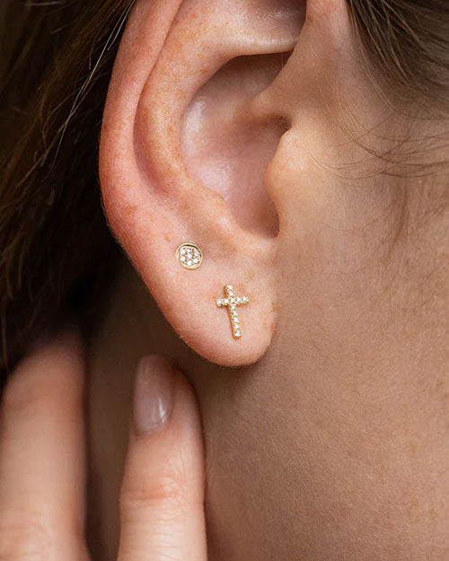 Close-up of an ear wearing two gold stud earrings: one small round stud with clear gemstones and one cross-shaped stud encrusted with small clear stones.