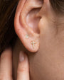 Close-up of an ear wearing two gold stud earrings: one small round stud with clear gemstones and one cross-shaped stud encrusted with small clear stones.