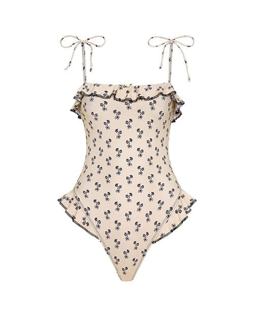 One-piece swimsuit in a cream color with a small black floral print pattern. The design features thin shoulder tie straps, ruffled trim along the neckline and hips, and a matching headscarf accessory.