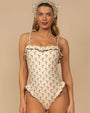 One-piece swimsuit in a cream color with a small black floral print pattern. The design features thin shoulder tie straps, ruffled trim along the neckline and hips, and a matching headscarf accessory.