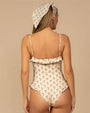 Back view of a one-piece swimsuit in a cream color with a small black floral print pattern. The design features thin shoulder tie straps, ruffled trim along the neckline and hips, and a matching headscarf accessory.