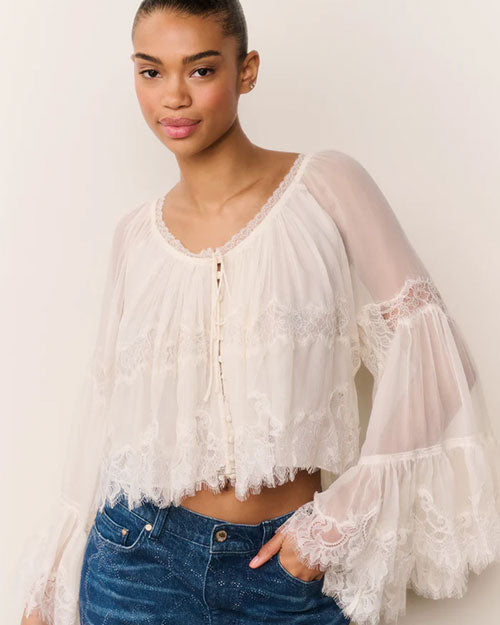 White sheer lace blouse with long flared sleeves, delicate lace trim, and front tie closure, paired with blue denim jeans, shown against a plain beige background.
