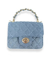 Small quilted handbag made of light blue denim fabric with a diamond stitch pattern, featuring a gold-tone twist-lock closure on the front flap and a short handle with interwoven blue material and gold chain links.