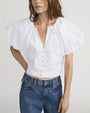 Person wearing a white short-sleeve blouse with voluminous puff sleeves and a deep V-neckline, featuring a row of small gold buttons down the front. The blouse is paired with high-waisted blue jeans.