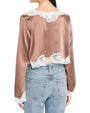 Back view of a blush satin cropped blouse with white scalloped lace trim at the neckline, hem, and cuffs, paired with light-wash jeans.