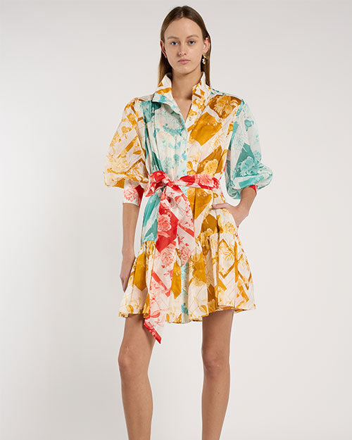 Person standing in a studio against a light neutral background, wearing a short, collared shirt dress with elbow-length sleeves and a tied waist sash. The dress features an abstract print in teal, mustard yellow, coral, and white. 