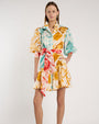 Person standing in a studio against a light neutral background, wearing a short, collared shirt dress with elbow-length sleeves and a tied waist sash. The dress features an abstract print in teal, mustard yellow, coral, and white. 