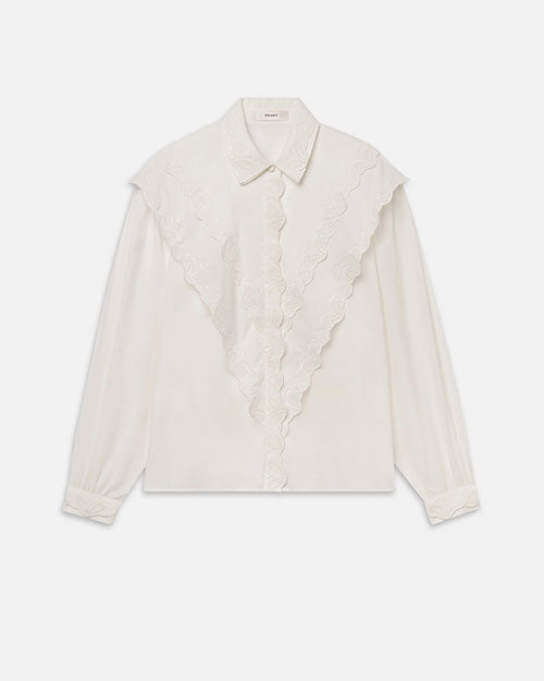 Front view of a white long-sleeve blouse displayed against a light gray background. The blouse features a pointed collar, concealed front button placket, and ornate scalloped ruffle detailing embroidered with floral motifs that forms a dramatic V-shape across the front from the shoulders to the center. The sleeves have a relaxed, slightly billowy fit and gather at buttoned cuffs, creating a soft, romantic silhouette.