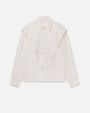 Front view of a white long-sleeve blouse displayed against a light gray background. The blouse features a pointed collar, concealed front button placket, and ornate scalloped ruffle detailing embroidered with floral motifs that forms a dramatic V-shape across the front from the shoulders to the center. The sleeves have a relaxed, slightly billowy fit and gather at buttoned cuffs, creating a soft, romantic silhouette.