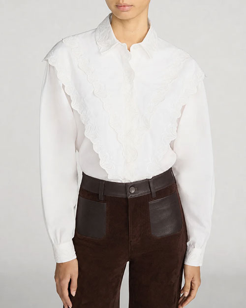 Front view of a white long-sleeve blouse with a pointed collar and intricate scalloped ruffle detailing that extends from the shoulders down the front on both sides of the hidden button placket. The blouse features a lightweight, slightly sheer fabric and gathered cuffs. It is paired with high-waisted dark brown pants featuring large patch pockets with contrasting leather-look panels and a button closure. The outfit is shown against a light gray background.
