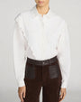 Front view of a white long-sleeve blouse with a pointed collar and intricate scalloped ruffle detailing that extends from the shoulders down the front on both sides of the hidden button placket. The blouse features a lightweight, slightly sheer fabric and gathered cuffs. It is paired with high-waisted dark brown pants featuring large patch pockets with contrasting leather-look panels and a button closure. The outfit is shown against a light gray background.