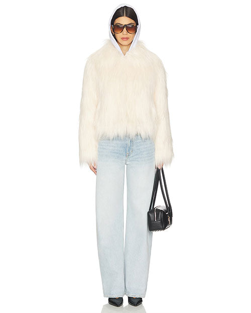Full-body view of a person wearing a white faux fur jacket with a shaggy texture, paired with light blue wide-leg jeans. The outfit is accessorized with a small black handbag held in the left hand and black pointed-toe shoes. The jacket has a slightly cropped length, and the overall look is casual yet stylish.
