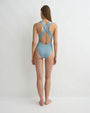 Back view of a person wearing a light grey blue one piece swimsuit with wide straps that crisscross across the back. The person is standing against a white background. 