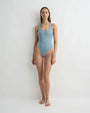 A person wearing a light grey blue one piece swimsuit with wide straps and standing in front of a white background. 