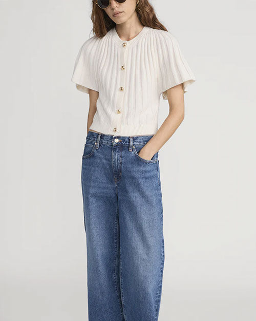 Person wearing a short-sleeve, cream-colored ribbed knit cardigan with gold buttons down the front, paired with high-waisted blue jeans. The cardigan has a structured, cape-like sleeve design.