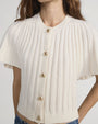 Close-up of a cream-colored ribbed knit short-sleeve cardigan featuring a round neckline and four gold buttons down the front. The fabric has a textured vertical rib pattern.