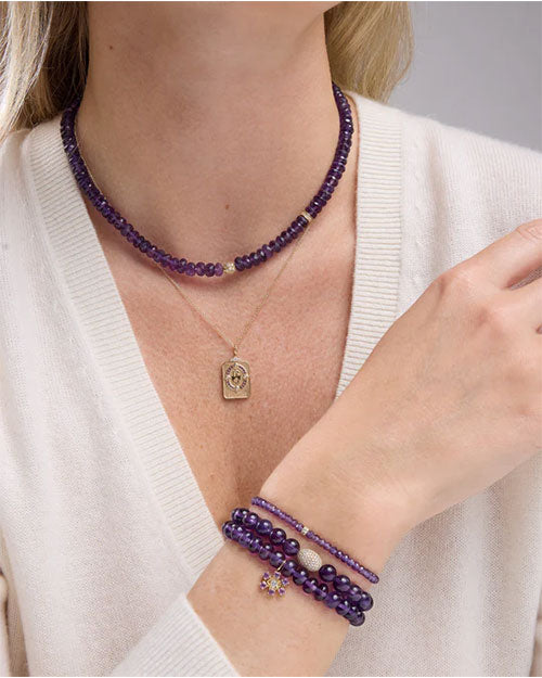 Person wearing a purple beaded necklace, a gold pendant necklace, and two matching purple beaded bracelets, paired with a white top.