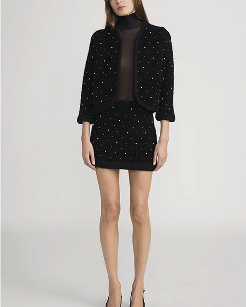 Model wearing a black textured two-piece outfit consisting of a cropped jacket and a mini skirt, both embellished with small pearl-like beads. The jacket has three-quarter sleeves and an open front, paired with a sheer black top underneath.