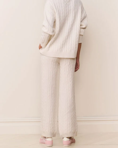 Back view of a cream-colored cable-knit sweater paired with matching wide-leg knit pants. Both pieces feature a textured cable pattern and a relaxed fit. The outfit is styled with light pink sneakers.