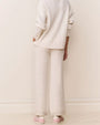 Back view of a cream-colored cable-knit sweater paired with matching wide-leg knit pants. Both pieces feature a textured cable pattern and a relaxed fit. The outfit is styled with light pink sneakers.