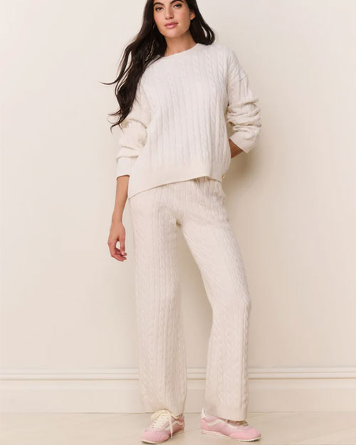 Front view of a cream-colored cable-knit sweater paired with matching wide-leg knit pants. The sweater has a relaxed fit with long sleeves and a ribbed crew neckline, while the pants feature the same textured cable pattern. The outfit is styled with light pink sneakers.