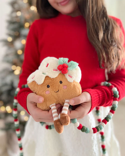  A person wearing a red long-sleeve top and a white textured skirt is holding a plush holiday-themed toy shaped like a festive treat. The toy has a light brown base, white icing with colorful sprinkles, a green felt holly leaf with red berries on top, and two small legs with striped stockings and green shoes. A garland made of red, green, and white felt balls is draped around the person’s arms, and a blurred Christmas tree with lights is visible in the background.