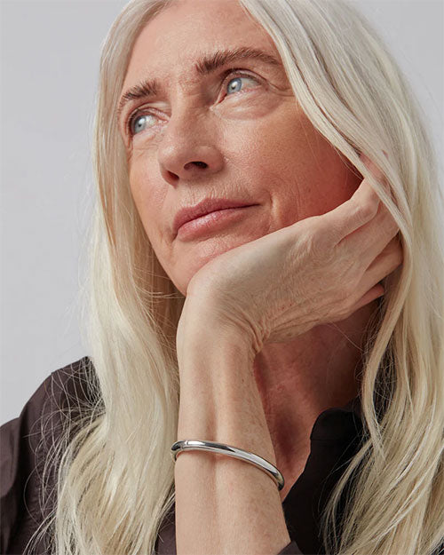 Close-up of a wrist wearing a sleek, polished silver bracelet with a minimalist design, paired with long light-colored hair and a dark sleeve.