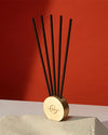 A minimalist gold stand with a circular base and five thin, elongated black stems protruding vertically from the center. The stand is placed against a red background on top of a concrete surface.