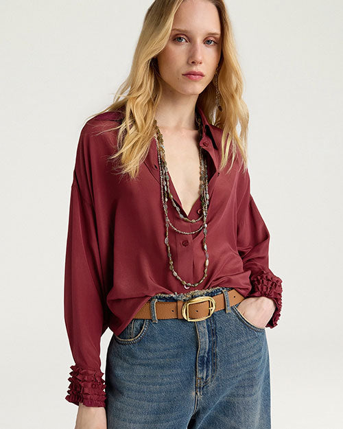 Close-up front view of a burgundy button-front blouse with a pointed collar and long sleeves featuring ruffled cuffs. The blouse is styled with blue denim jeans, a tan leather belt with a gold-tone buckle, and layered beaded necklaces. Shown against a light gray background.