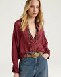 Close-up front view of a burgundy button-front blouse with a pointed collar and long sleeves featuring ruffled cuffs. The blouse is styled with blue denim jeans, a tan leather belt with a gold-tone buckle, and layered beaded necklaces. Shown against a light gray background.