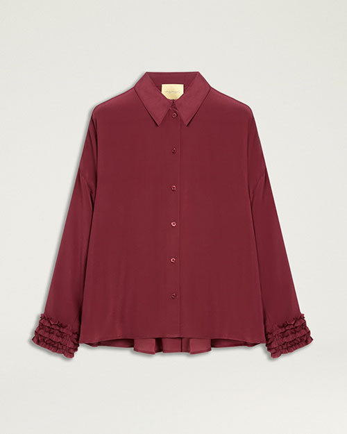 Product image of a burgundy button-front blouse with a pointed collar, long sleeves, and a relaxed silhouette. The blouse features ruffled cuff detailing, tonal buttons, and a slightly curved hem with an extended back panel. Shown laid flat against a light gray background.