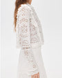 A person wearing a white long‑sleeve eyelet lace blouse shown from the side. The blouse features intricate cutout embroidery in floral and geometric patterns, ruffled trim along the shoulders, and flared sleeves with lace edging. The hem has delicate scalloped details. The person is also wearing a white pleated skirt, and the background is plain and light-colored.