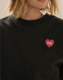 Close-up view of a black crewneck sweatshirt with a small red heart graphic with white text on the upper left chest.