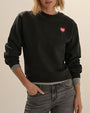 Close-up view of a black crewneck sweatshirt with long sleeves and ribbed cuffs and hem. The sweatshirt features a small red heart graphic with white text on the upper left chest and is styled with gray denim jeans and a gray layer visible at the cuffs and hem. Shown against a light neutral background.