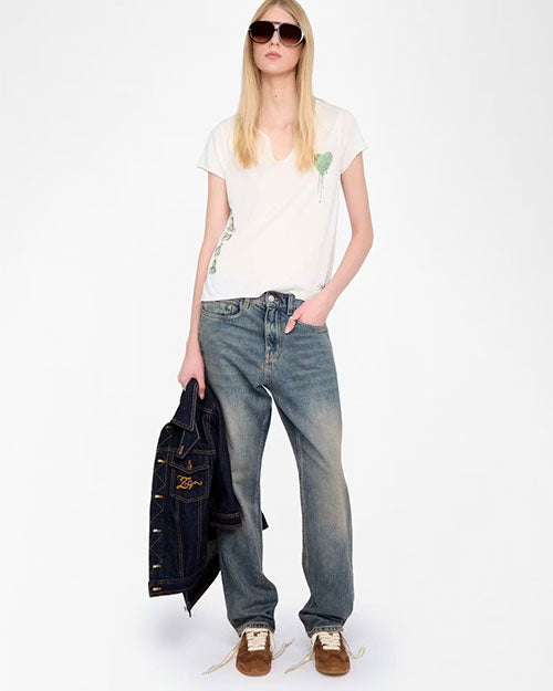 A person is standing against a plain white background wearing a casual outfit consisting of a white short-sleeve T-shirt with a small green graphic on the chest, loose-fitting faded blue jeans, and brown sneakers with white laces. The person is holding a dark blue denim jacket with visible stitching and a logo on the left side in their left hand, while their right hand is placed in the front pocket of the jeans.