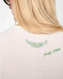 Close-up of the back of a white T-shirt featuring decorative green rhinestone designs. The embellishments include a wing-like shape near the upper back and a smaller curved line of rhinestones below it. The person has long, straight blonde hair partially covering the top of the shirt.