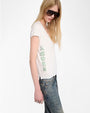 A person is shown from the side wearing a white short-sleeve T-shirt with the word “AMOUR” printed vertically in large green letters along the left side. The outfit also includes faded blue jeans and a silver ring on the left hand. The background is plain white.