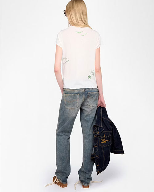  A person is standing against a plain white background, facing away from the camera. The outfit includes a white short-sleeve T-shirt with small green graphics and text on the back, faded blue straight-leg jeans, and brown sneakers with white laces. The person is holding a dark blue denim jacket with visible stitching and a logo on the back pocket area in their left hand.