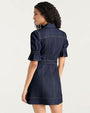 Back three-quarter view of a dark denim mini dress with short puff sleeves, contrast topstitching, a fitted waist with belt, and seam detailing across the back, photographed against a light studio background.