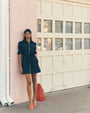 Full-body view of a person wearing a dark denim mini dress with short sleeves, front zipper, contrast stitching, and a cinched waist, standing on a sidewalk and leaning against a pale pink exterior wall near a white garage door, with orange heeled sandals visible.