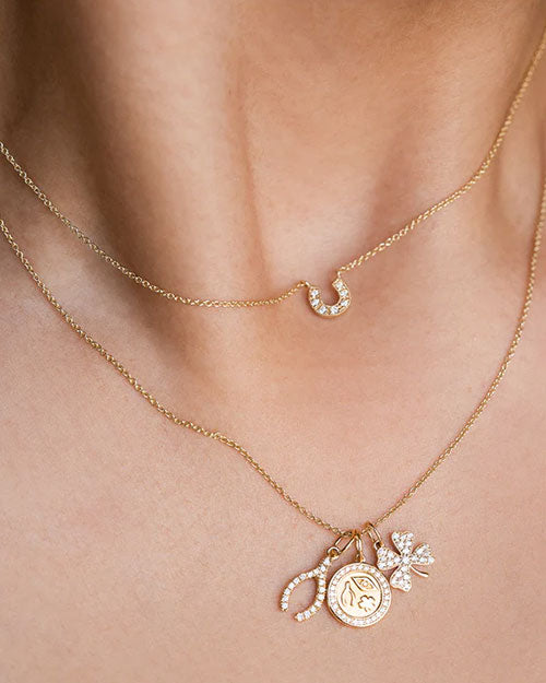 Two layered gold necklaces with delicate chains. The upper necklace has a diamond-studded horseshoe pendant, while the lower necklace features multiple charms, including a horseshoe, an eye medallion, and a four-leaf clover, all adorned with small diamonds.