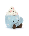 Plush toy shaped like a blue mug with embroidered snowflakes, a smiling face, and two brown legs. The top features white plush whipped cream with pink accents and brown sprinkles, resembling a cozy hot beverage.