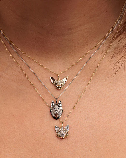 Close-up of a person wearing three layered necklaces with dog-shaped pendants. The top pendant is a small gold Chihuahua head, the middle pendant is a silver Husky head with dark accents, and the bottom pendant is a gold French Bulldog head. Each pendant is detailed with sparkling stones and attached to delicate chains in gold and silver tones.