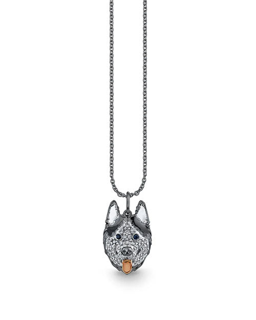 A silver-toned necklace featuring a pendant shaped like a husky dog’s head. The pendant is encrusted with small, sparkling stones, has dark blue gemstone eyes, and a rose-gold colored tongue detail. The chain is a simple, fine link design.
