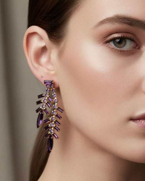 Close-up of an ear wearing a gold-toned statement earring featuring cascading branches of purple baguette-cut crystals and marquise-shaped stones, accented with small round clear crystals.