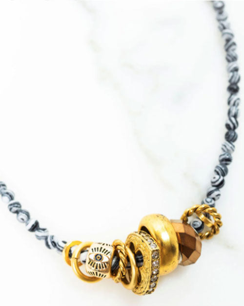 Close-up of a necklace with black and white patterned beads and a cluster of metallic charms at the center, including multiple gold rings, a bronze faceted bead, and a silver charm with engraved details, displayed on a white marble surface.
